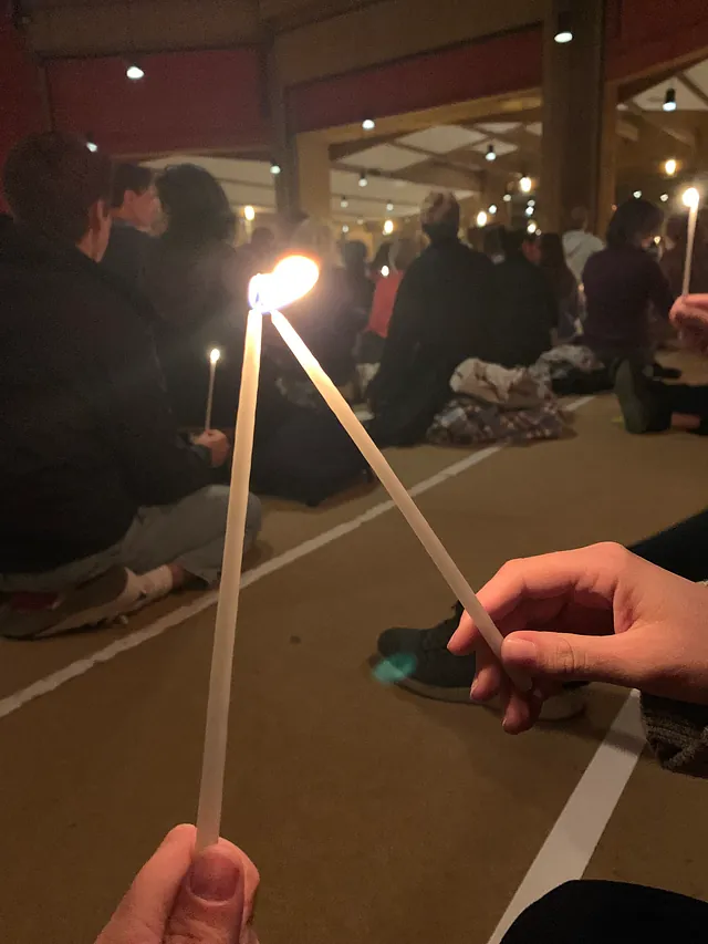 Menschen sitzen in einer großen Halle im Kerzenlicht; zwei Personen entzünden ihre Kerzen aneinander, um das Licht weiterzugeben