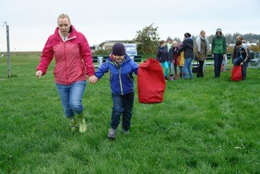 Hier sieht man ein Kind und eine Frau auf dem Rasen laufen. Im Hintergrund stehen noch mehr Menschen. Alle tragen eine Regenjacke. Der Himmel ist leicht bewölkt.