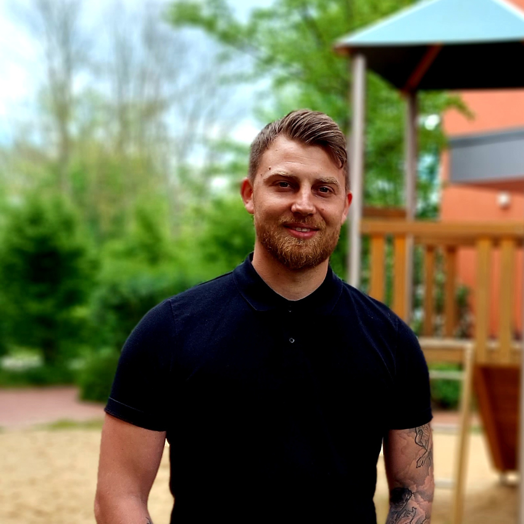 Auf diesem Bild sieht man einen jungen Erwachsenen Herrn, welcher dunkel Braune Haare hat und ein schwarzes T-Shirt trägt. Er hat Tatoos am rechten Arm. Er sticht aus dem Foto heraus da der Hintergrund verschwommen ist. Im Hintergrund kann man grob Bäume und Büsche und einen Spielplatzt erkennen.