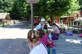 Auf diesem Bild erkennt man, dass die Kinder in einem Freizeitpark sind. Im HIntergrund sind ganz viele Kinder die auf Spielplätzen spielen. Im Vordergrund sind Kinder die in ihren Taschen wühlen oder etwas essen. Ein Mädchen guckt lächelnd in die Kamera und hat eine weiße Mütze auf.
